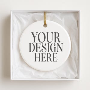 May include: A white ceramic ornament with the text "YOUR DESIGN HERE" in dark gray, presented in a white gift box with tissue paper. The ornament has a gold string for hanging.