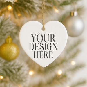 May include: A white heart-shaped Christmas ornament with the text "YOUR DESIGN HERE" in dark gray. The ornament hangs from a gold string, with a blurred background of a Christmas tree and other ornaments.