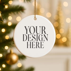 May include: A white, round Christmas ornament with the text "YOUR DESIGN HERE" in black. The ornament hangs from a gold string, with a blurred background of Christmas lights and a decorated tree.