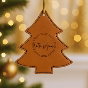 Puede incluir: Adorno navideño de cuero marrón con forma de árbol, con costuras negras y una cuerda dorada para colgar. El texto "Little Mocks" está dentro de un círculo. El fondo está borroso con luces bokeh.