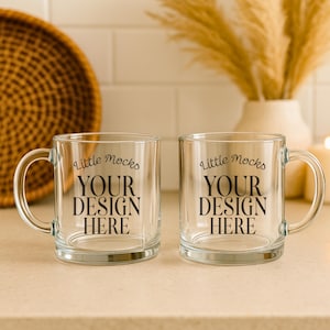 May include: Two clear glass mugs with handles. Each mug has the text "Little Mocks YOUR DESIGN HERE" in black. The mugs are on a light-coloured surface.