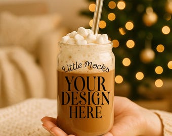 Puede incluir: Un tarro de cristal lleno de una bebida marrón, cubierto con espuma blanca y malvaviscos, y una pajita transparente. El tarro tiene el texto "Little Mocks" y "YOUR DESIGN HERE". El fondo incluye un árbol de Navidad con luces borrosas.