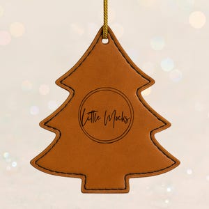 Puede incluir: Adorno navideño de cuero marrón con costuras negras y una cuerda dorada para colgar. El adorno tiene el texto "Little Mocks" dentro de un círculo en el centro.