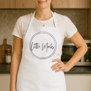 May include: White apron with the text "Little Mocks" in a black, handwritten-style font, encircled by a double black line. The apron is being worn in a kitchen setting.