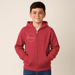 May include: A red zip-up hoodie with a hood and pockets. The hoodie has the text "Little Mocks" in a white circular design on the front. The model is wearing blue jeans.