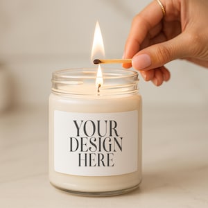 以下が含まれることがあります： 「YOUR DESIGN HERE」と書かれた白いラベルが付いた、透明なガラス瓶に入った点灯したキャンドル。 炎のついたマッチが芯の上に保持されています。 キャンドルは明るい色の表面にあります。