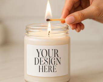May include: A lit candle in a clear glass jar with a white label that reads "YOUR DESIGN HERE". A match is held above the wick, with a flame lit. The candle is on a light-colored surface.