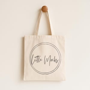 May include: A cream-colored tote bag with the text "Little Mocks" in a handwritten style within a double circle. The bag hangs from a wooden peg on a light-colored wall. The bag has long handles.