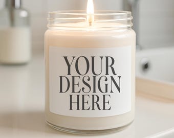 May include: A lit candle in a clear glass jar with a white label that reads "YOUR DESIGN HERE". The candle has a cream-colored wax and is sitting on a white surface. The background is blurred, showing a bathroom setting.