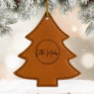 Puede incluir: Adorno navideño de cuero marrón con forma de árbol, con costuras negras y una cuerda dorada para colgar. El adorno tiene el texto "Little Mocks" en un diseño circular. El fondo está borroso con ramas nevadas.