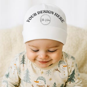May include: A white baby beanie with the text "YOUR DESIGN HERE" in gray. The beanie is worn by a baby wearing a beige and light blue patterned onesie with snowmen and trees.