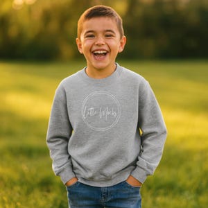 May include: A gray sweatshirt with the text "Little Mocks" inside a white circle. The sweatshirt is worn by a child who is smiling. The child is wearing blue jeans and is standing in a grassy field.