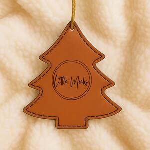 Puede incluir: Adorno navideño de cuero marrón con forma de árbol, bordes cosidos y una cuerda dorada para colgar. El adorno tiene el texto "Little Mocks" dentro de un círculo.