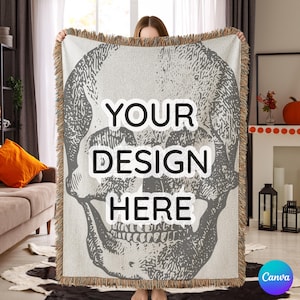 May include: A light gray throw blanket featuring a large skull design. The blanket has a fringed edge and the text "YOUR DESIGN HERE" in a bold font. The blanket is held up in front of a living room setting.