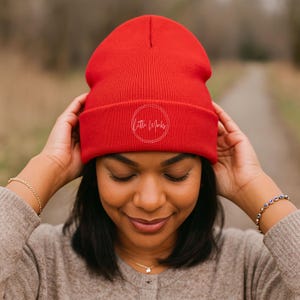 May include: A bright red beanie with the text "Little Mucks" in a white circle. The beanie is made of a soft, knitted material. The person is wearing a tan sweater and gold bracelets.
