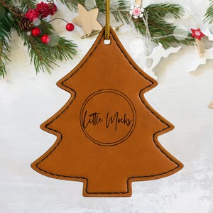 Puede incluir: Adorno navideño de cuero marrón con forma de árbol, con el texto "Little Mocks" en un círculo. Tiene un borde cosido y una cuerda dorada para colgar. Decoración festiva navideña.