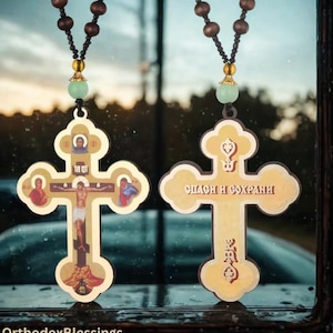 May include: Two wooden Orthodox crosses with religious iconography, suspended by beaded strings. The crosses are gold-toned with detailed images of Jesus and saints. One cross has the text "TA OT" and the other has text in Cyrillic script. The background is blurred.