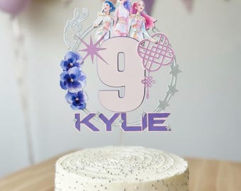 May include: A white frosted cake on a white cake stand, topped with a decorative topper. The topper features a number 9, the name "Kylie", and illustrations of three figures. The cake is decorated with silver sprinkles.
