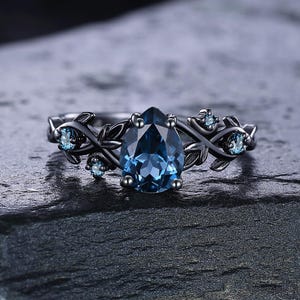 Könnte beinhalten: Ein schwarzer Ring mit einem birnenförmigen, blauen Edelstein als Mittelpunkt. Der Ring hat ein florales Design mit kleineren blauen Edelsteinen und Blattakzenten. Der Ring liegt auf einer dunklen, strukturierten Oberfläche.