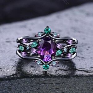 Puede incluir: Un anillo de metal negro con una gran piedra púrpura de corte ovalado en el centro, rodeada de piedras púrpuras y turquesas más pequeñas. El anillo tiene un diseño único y en capas con detalles intrincados, sobre un fondo oscuro y texturizado.