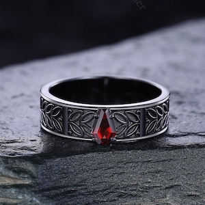 Puede incluir: Un anillo plateado con un diseño geométrico y una piedra preciosa roja. El anillo presenta un intrincado patrón de hojas y líneas alrededor de la banda. La piedra preciosa es de color rojo vibrante y tiene una forma única.