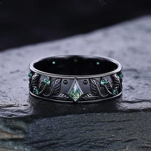Könnte beinhalten: Schwarzer Ring mit einem von der Natur inspirierten Design. Er hat einen zentralen, hellgrünen, rautenförmigen Stein und kleinere grüne Steine. Das Band ist mit Blatt- und Rankenmustern verziert. Der Ring liegt auf einer dunklen, strukturierten Oberfläche.