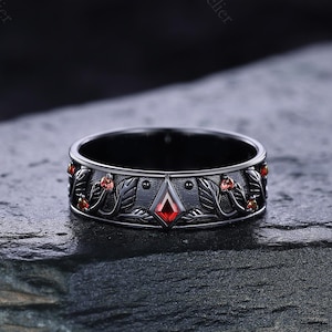 May include: A black ring with a detailed floral design. The ring features red gemstones and a central red diamond-shaped stone. The band is dark, with intricate leaf and berry patterns. The ring is displayed on a dark, textured surface.