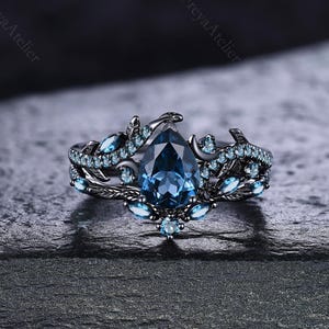 May include: A dark, ornate ring with a large, pear-shaped blue gemstone as the focal point. The ring features a detailed, floral-inspired design with smaller blue gemstones and dark metal accents. The ring is set against a dark, textured background.
