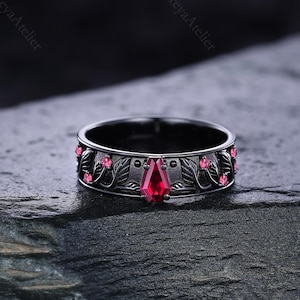 May include: Black ring with a central, teardrop-shaped red gemstone. The ring features a floral and leaf design with small pink gemstones. The ring is set on a dark, textured surface.