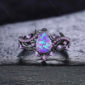 May include: A black metal ring featuring a central pear-shaped purple opal stone surrounded by small, marquise-cut and round-cut purple accent stones. The band is designed with a vine-like motif that wraps around the central setting.
