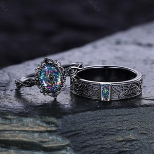 May include: Two black rings with iridescent stones. One ring features an oval-shaped stone with a decorative filigree setting. The other ring has a band with a floral pattern and a rectangular iridescent stone. The rings are displayed on a dark, textured surface.