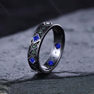 Puede incluir: Un anillo gris oscuro con un patrón repetitivo de piedras preciosas cuadradas azules y redondas verdes. El anillo tiene un diseño intrincado y un acabado pulido. El anillo está sobre una superficie de piedra gris oscura.