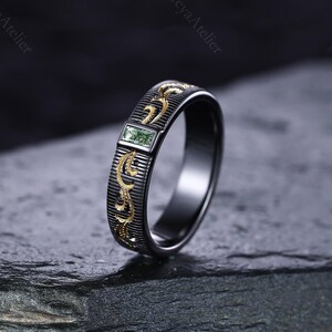 May include: A black ring with gold filigree and a rectangular green gemstone. The ring has a dark, textured band with vertical lines and gold floral designs. The gemstone is set in a rectangular gold frame.