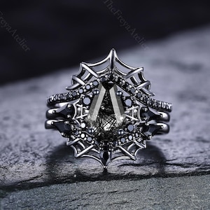May include: A three-piece black ring set with a unique design. The engagement ring features a large, triangular-cut black gemstone, surrounded by a spiderweb-like setting. The set includes two additional bands with smaller black stones.