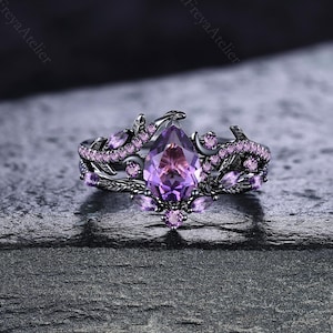 May include: A black ring with a large, pear-shaped purple gemstone at the center. The ring is adorned with smaller purple gemstones and a decorative, floral-inspired design. The ring is set against a dark, textured background.