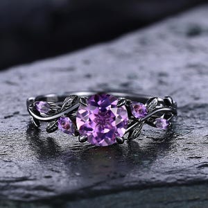 May include: A silver ring featuring a central, round, light purple gemstone, complemented by smaller purple stones and leaf detailing. The ring is set on a dark, textured surface.