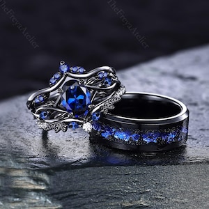 Könnte beinhalten: Ein Ring-Set aus schwarzem Metall, bestehend aus einem Verlobungsring mit einem ovalen blauen Mittelstein und blattartigen Verzierungen sowie einem passenden Ring mit einer blauen Einlage. Beide Ringe sind aus dunklem Metall gefertigt.