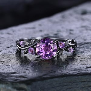 May include: A silver ring with a large, round, light purple gemstone at the center, flanked by smaller purple stones and leaf-shaped accents. The ring is set against a dark, textured background.