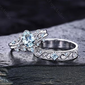 May include: A silver-toned engagement ring set featuring a central oval-cut light blue gemstone surrounded by small clear stones and leaf-like metal details, paired with a matching band that includes a square-cut light blue stone and scrollwork.