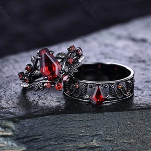 Peut inclure: Deux bagues noires ornées de pierres rouges. L'une présente une grosse pierre en forme de goutte et un motif floral. L'autre a une pierre plus petite en forme de goutte et une bande avec un motif de feuilles. Les bagues sont posées sur une surface sombre et texturée.