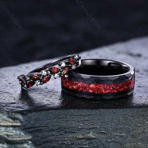 Puede incluir: Dos anillos negros con piedras preciosas rojas y blancas. Uno presenta una banda de piedras rojas, el otro una fila de piedras rojas y detalles blancos. Los anillos se exhiben sobre una superficie oscura y texturizada.