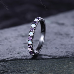 May include: A silver ring with a row of alternating purple and white gemstones. The ring is set against a dark, textured background, highlighting the sparkle of the stones. The Freya Atelier is printed on the top right and left of the image.