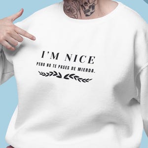 May include: White sweatshirt with the text "I'M NICE" and a Spanish phrase below. The sweatshirt has a crew neck and long sleeves. The person is wearing pink pants. The background is a mix of pink and blue shapes.