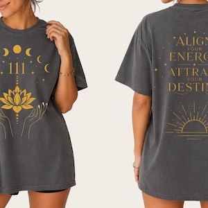 Puede incluir: Camiseta gris oscuro con diseño dorado. La parte delantera presenta una flor de loto, manos, lunas y el número 111. La parte trasera tiene el texto "ALIGN YOUR ENERGY ATTRACT YOUR DESTINY" con un gráfico de sol.