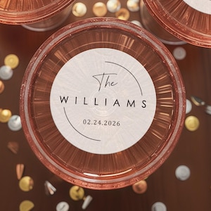 May include: Overhead view of three champagne glasses filled with a pink beverage. Each glass has a circular sticker with the text "The Williams" and the date "02.24.2026". The glasses are on a brown surface with scattered confetti.