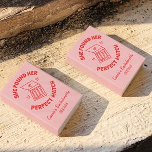 May include: Two pink matchboxes with the text "SHE FOUND HER PERFECT MATCH" in red. The boxes also include the text "Cassie's Bachelorette 06.12.2026" and a graphic of matches. The matchboxes are on a wooden surface.