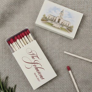 May include: Two rectangular matchboxes with white covers. One box is open, revealing wooden matches with dark red tips. The cover features the text The Johnsons and the date 06.06.2027 in red script. The second box shows a watercolor illustration of a building.