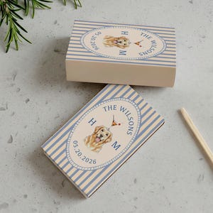 May include: Two matchboxes with a light blue and white striped design. Each box features a watercolor illustration of a golden retriever, the text "THE WILSONS", initials, and a date. Two matches with red tips are also visible.