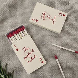 May include: Two rectangular matchboxes with cream-colored covers and red text. One box is open, revealing wooden matches with red tips. The text reads Jake & Ana, 6, 13, J, A, and The perfect match, accompanied by small red heart icons.