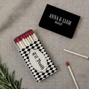 May include: Two rectangular matchboxes with red-tipped matches. One box is black with white text reading ANNA & LIAM 05.24.27. The other box features a black and white checkered pattern with an oval label reading 'til Death in black script.